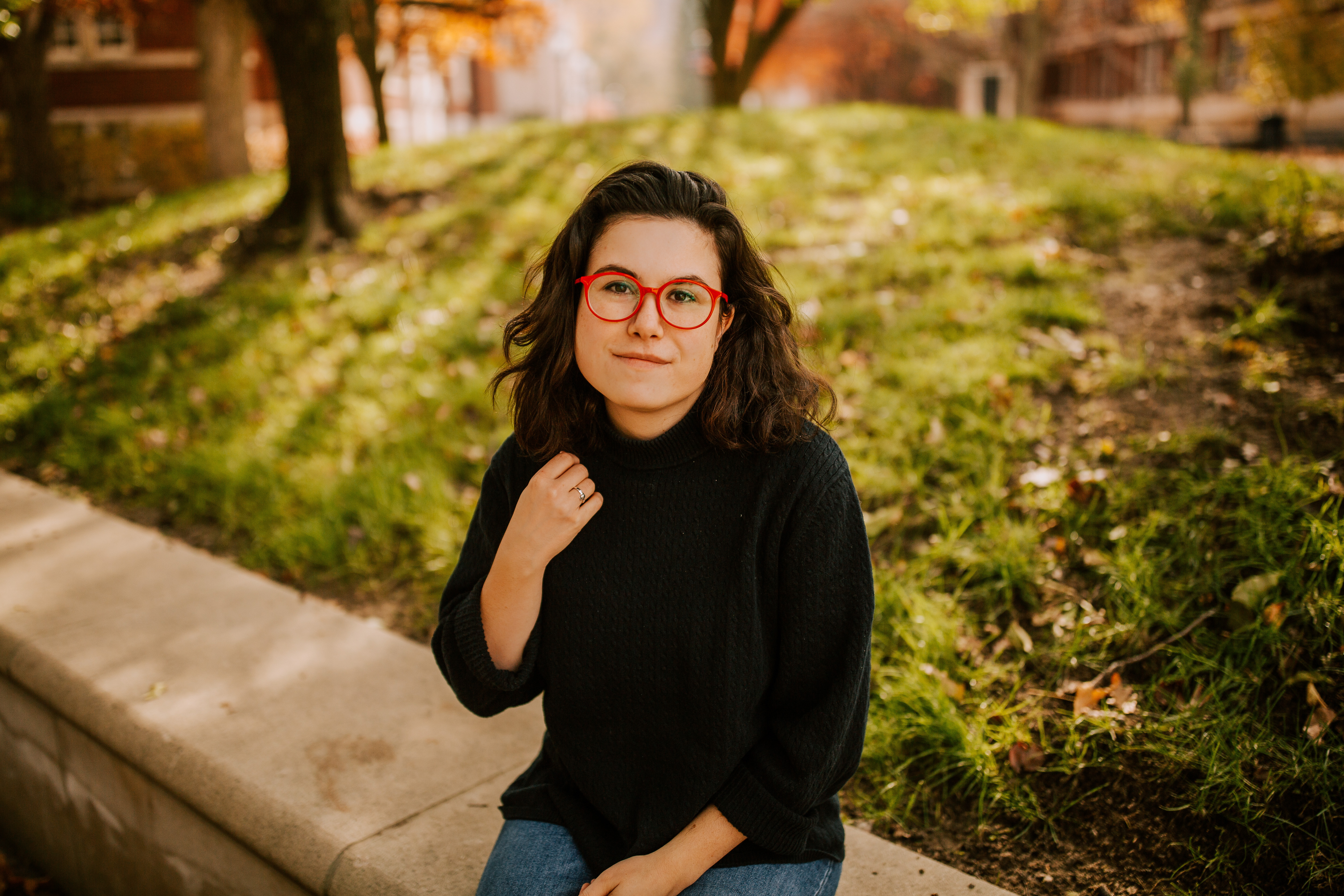 A photo of Kate O'Donoghue taken outdoors on a sunny day. Kate is dark-haired and wearing a black sweater. She had round red glasses.